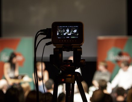 Im Hintergrund sind Personen auf einer Bühne bei einer Podiumsdiskussion zu sehen. Im Vordergrund eine Kamera, die aufzeichnet.